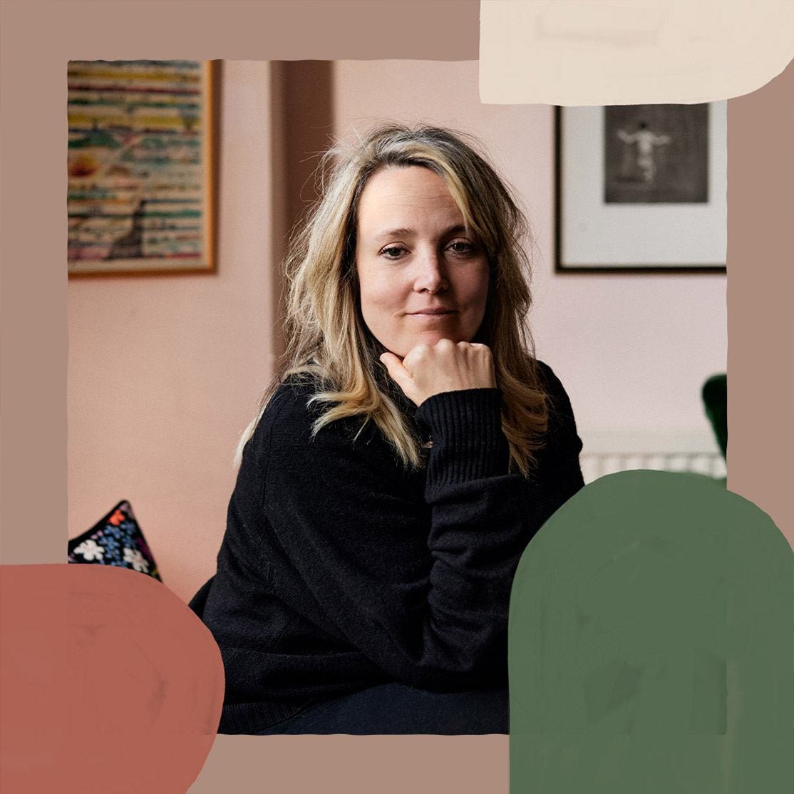 Bay Garnett is wearing a black jumper and is looking at the camera, smiling with her head resting on her hand. In the background, two picture frames are hung on a light pink wall.