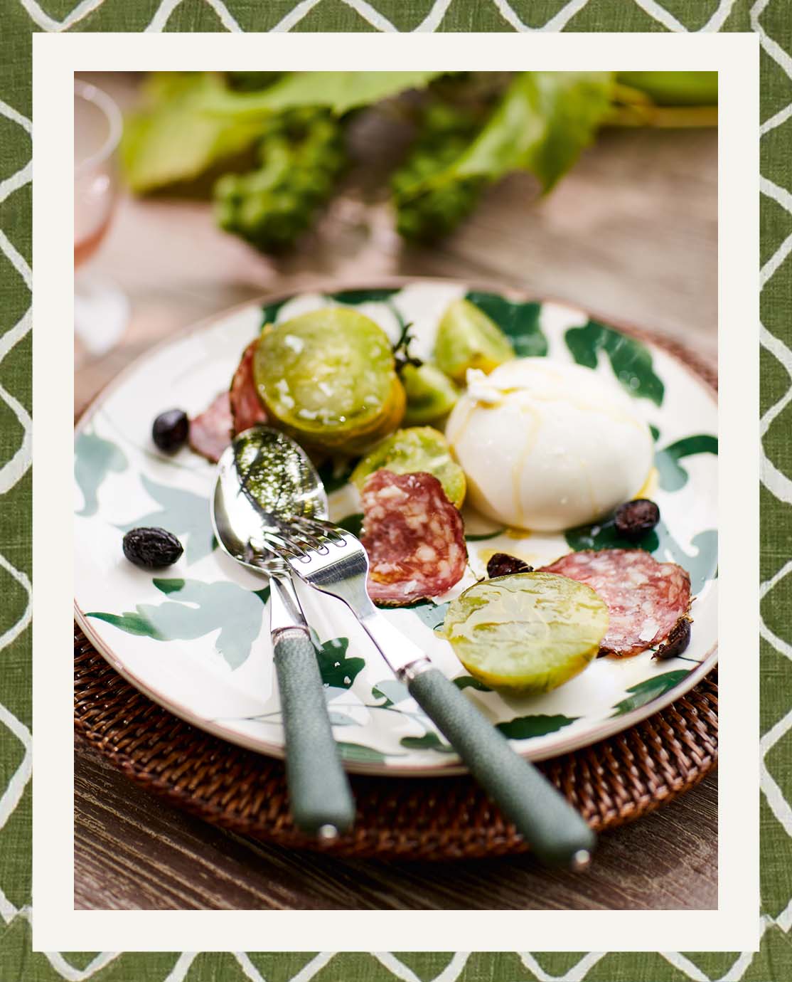 A green tomato, mozzarella and salami salad on a leaf-print plate. Faux shagreen knife and fork sit on top of it.
