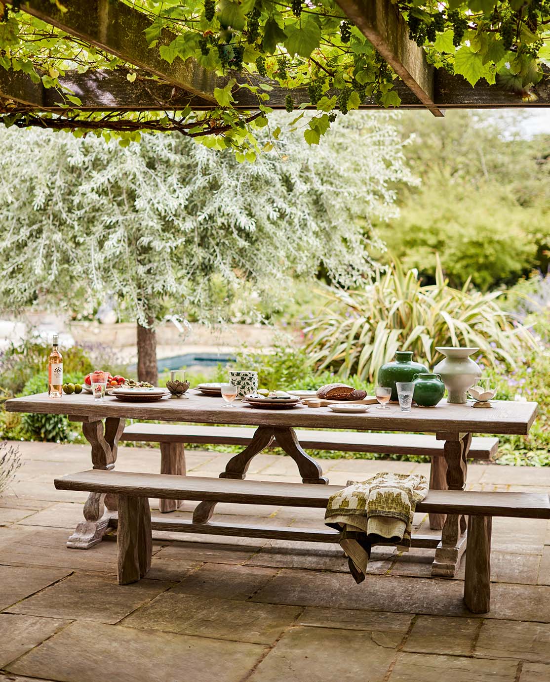 A large wooden dining table and matching wooden benches sit underneath a wooden canopy which is decorated with vines. The table is decorated with plates and colourful pots.