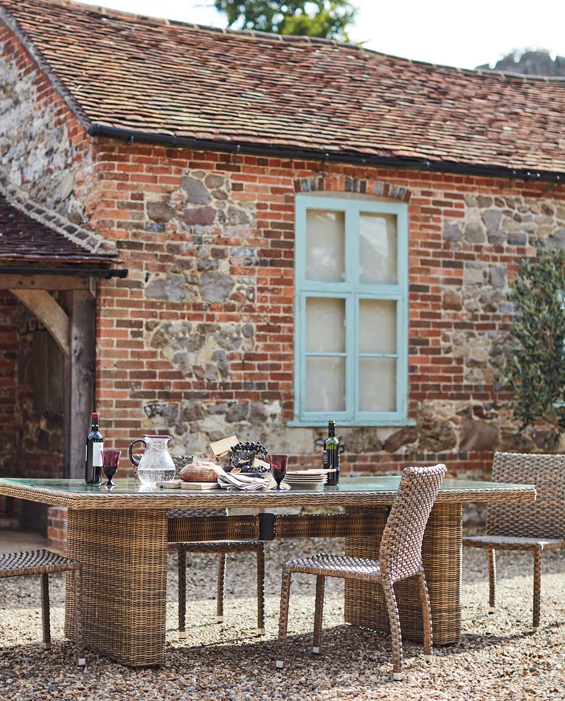 A rattan outdoor dining table and matching chairs sits in front of a red-brick house. It is decorated with plates, food and wine glasses.