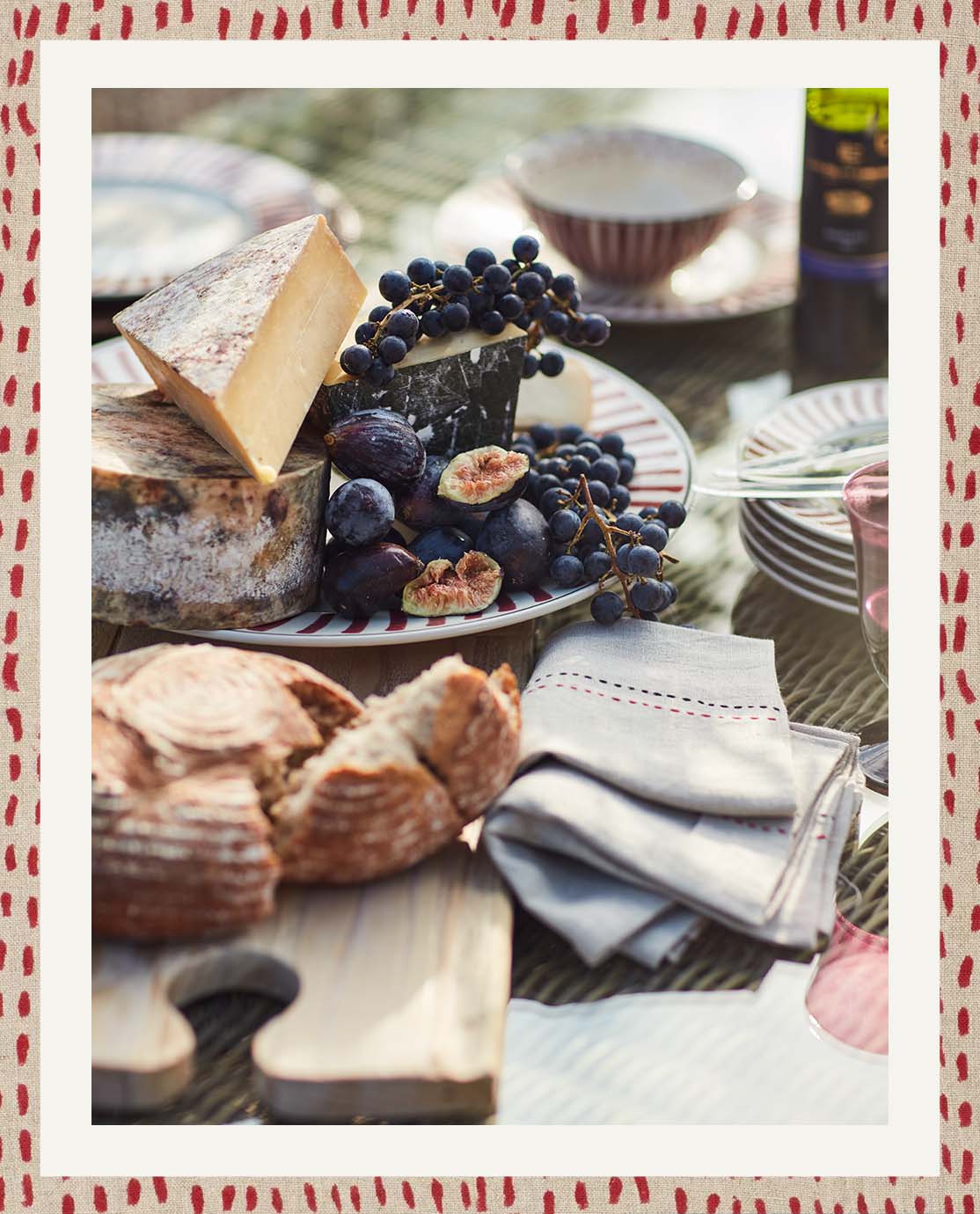 A close-up of food on an outdoor table; there is bread on a wooden cutting board, cheese, grapes and figs on a plate and napkins.