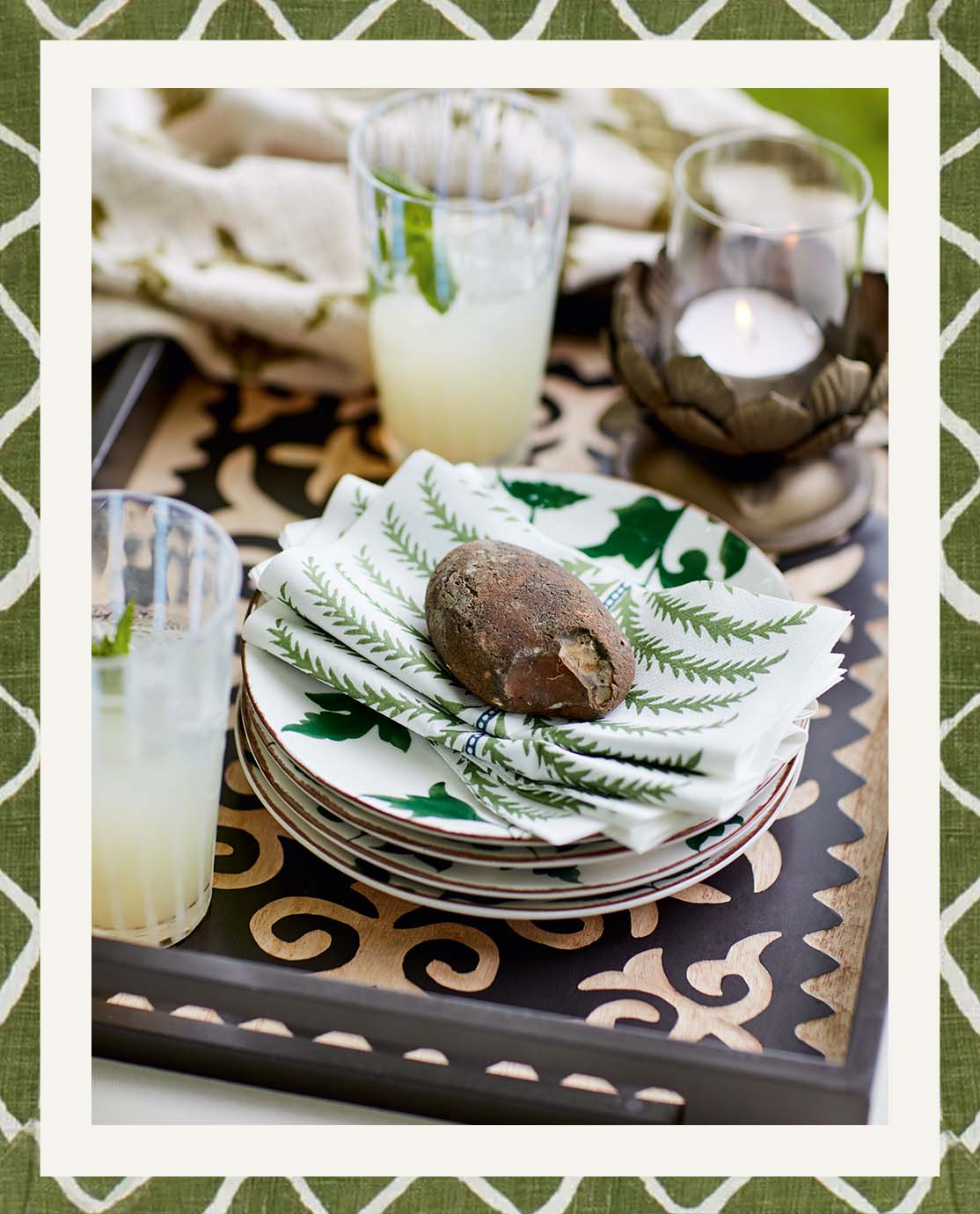 A wooden carved serving tray holds a stack of leaf=print plates, some palm-print napkins and two printed glasses holding cloudy lemonade.