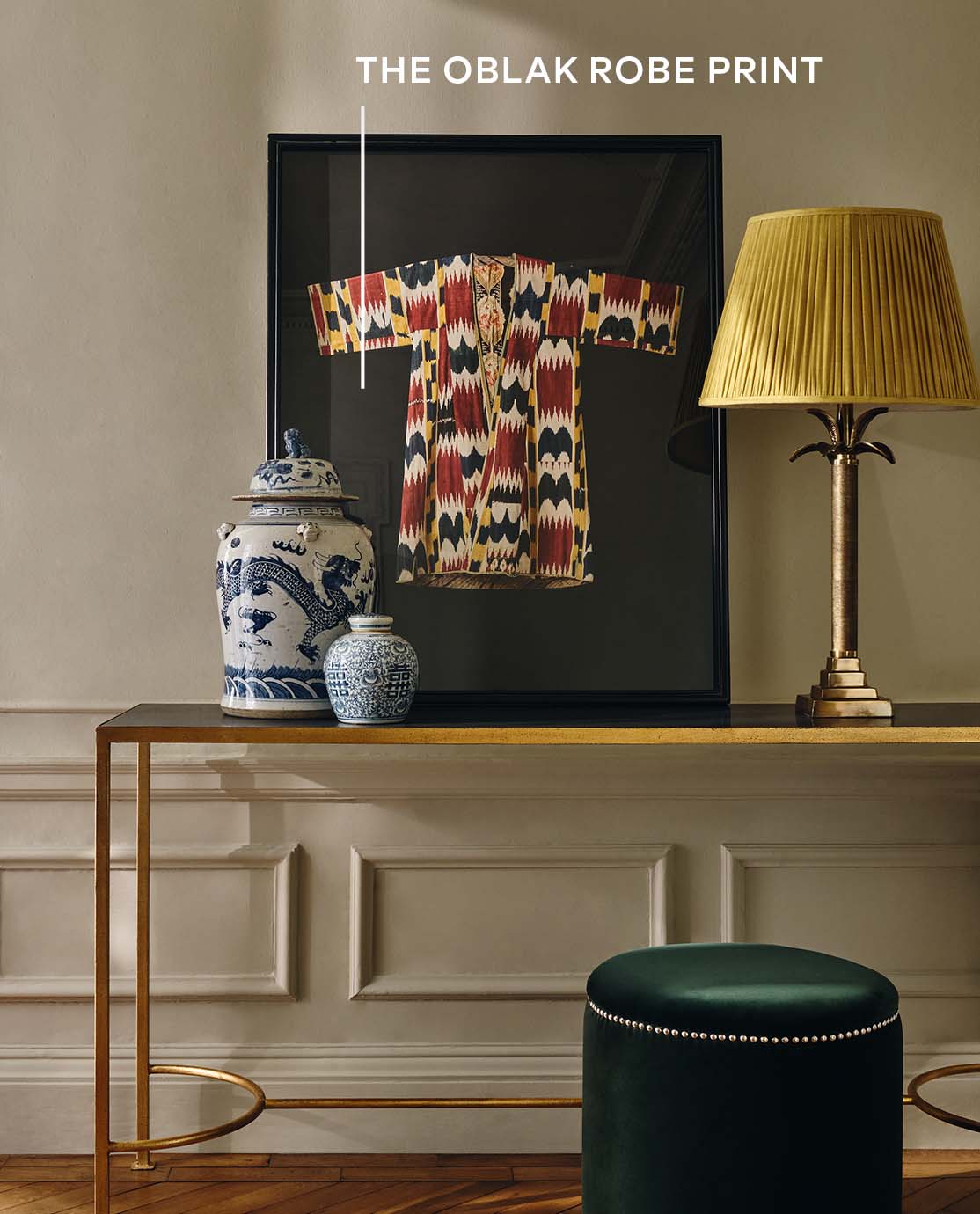 A console table with a yellow lamp, a robe print, ceramic pots and a green velvet footstool beside.