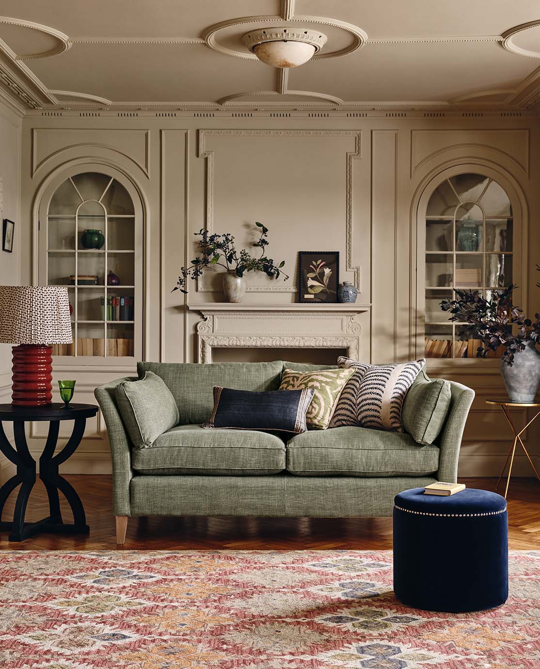 A country sitting room with a green linen sofa, a blue velvet footstool, a patterned rug, side table and red lamp placed in front of a fire place.