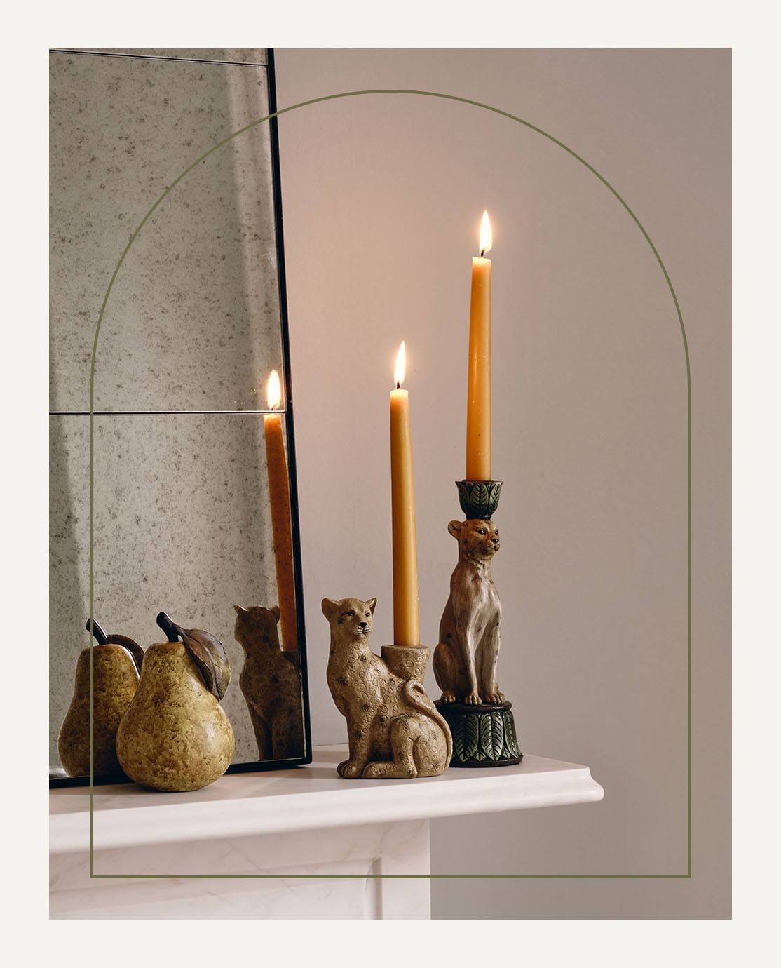 The corner of a mantel, decorated with leopard candle holders, a pear ornament and a mirror. 