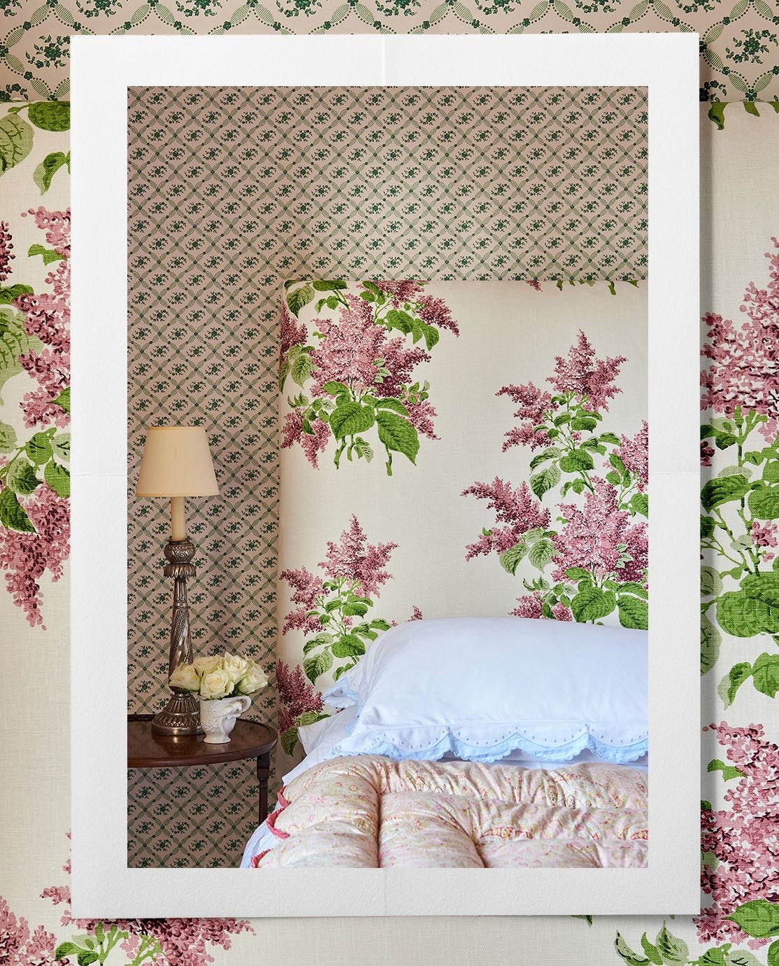 A bed decorated with a floral headboard and quilted bedding, next to a bedside lamp, in front of patterned wallpaper. 