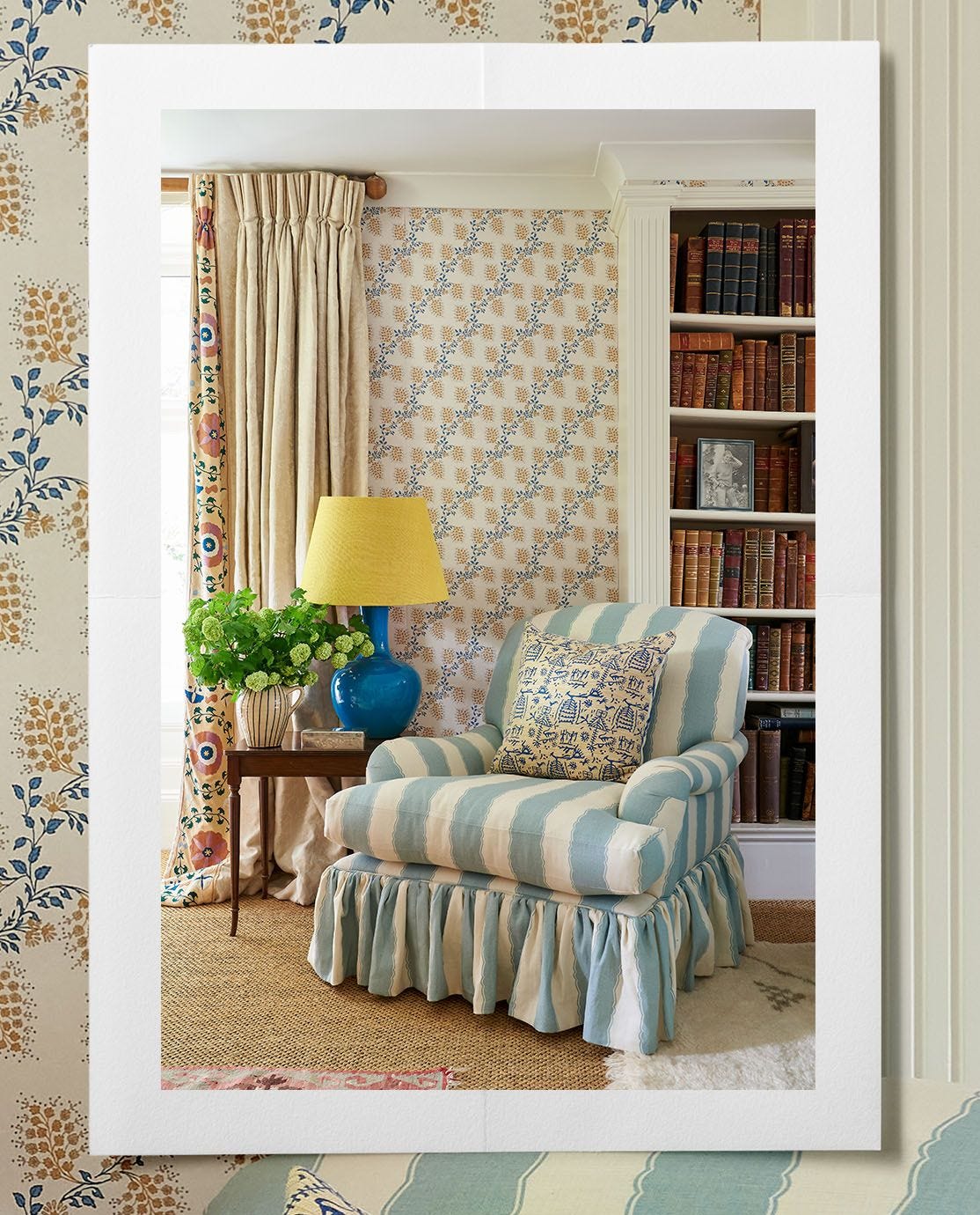 A corner of a sitting room featuring a blue striped armchair next to a side table with a lamp, behind a bookshelf, patterned wallpaper and long draped curtains. 