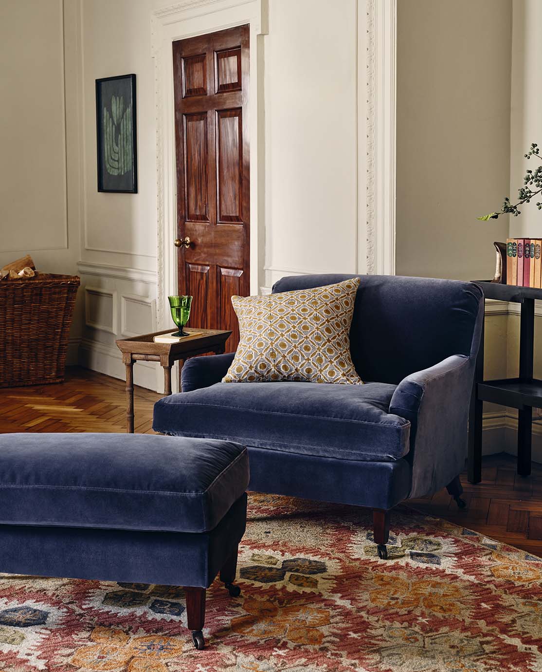A sitting room featuring a blue velvet armchair and a footstool with a patterned rug underneath. Behind is a wooden side-table and a dark wooden door.
