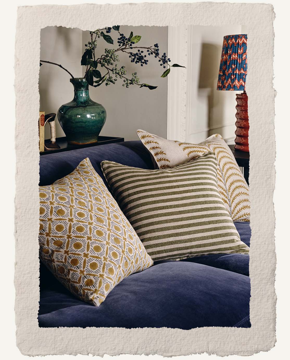 A close up of a blue velvet sofa decorated with patterned cushions in green and mustard yellow. In the background is a blue vase holding flowers.