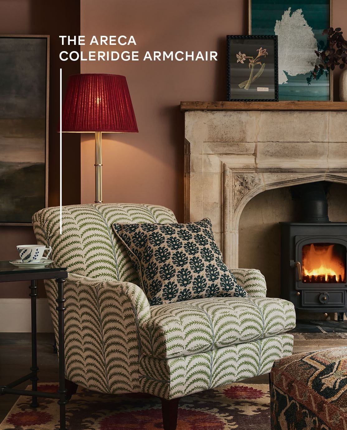 The corner of a cottage sitting room with a palm-print armchair in front a red lamp and a fireplace, decorated with framed art work.