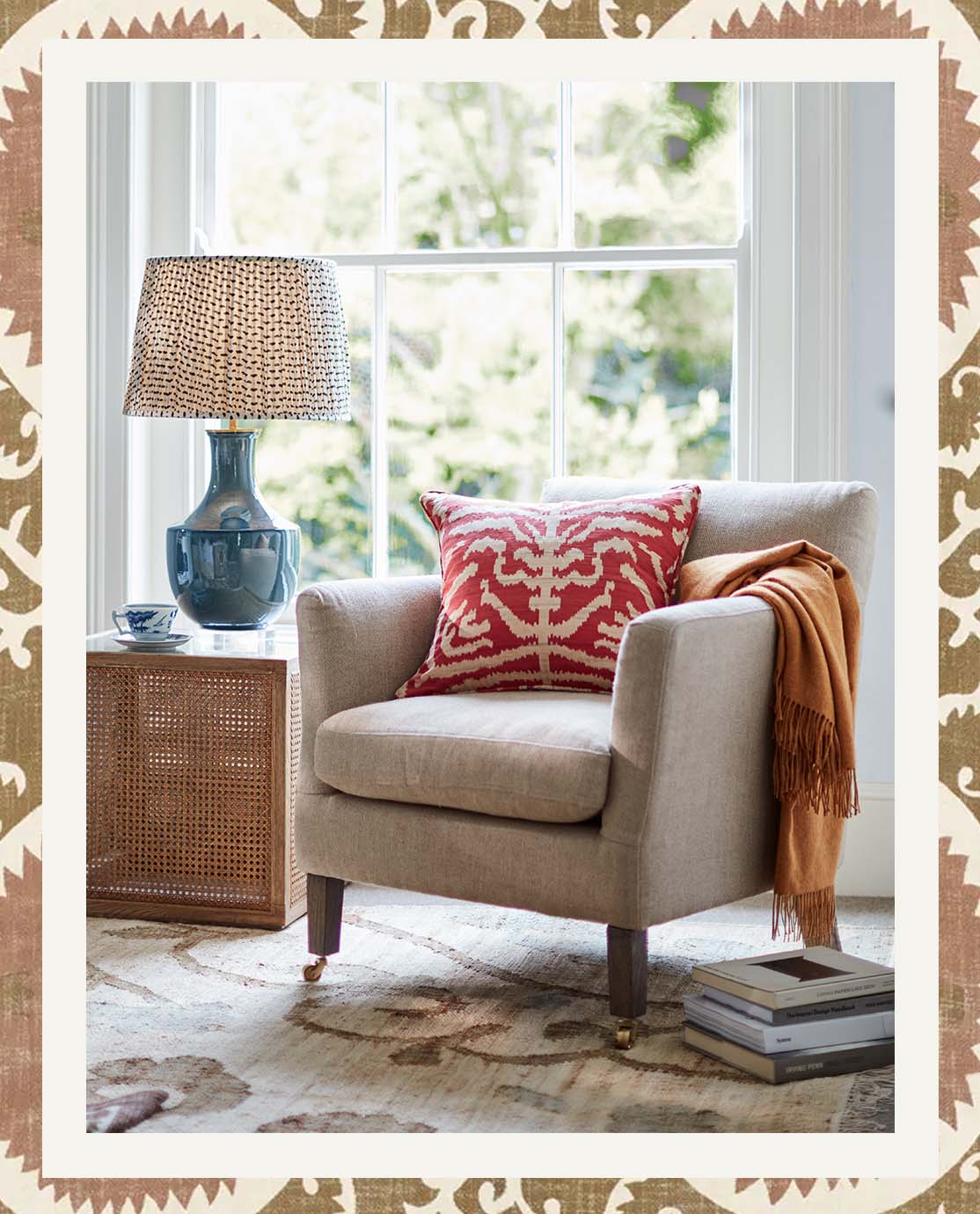 A linen armchair is decorated with a red printed pillow and orange throw. Next to it is a wooden side table and a blue lamp.