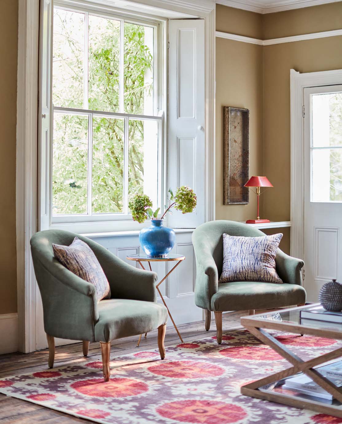 Two green armchairs sit on top of a red printed rug. A gold side table sits between them, decorated with a blue pot