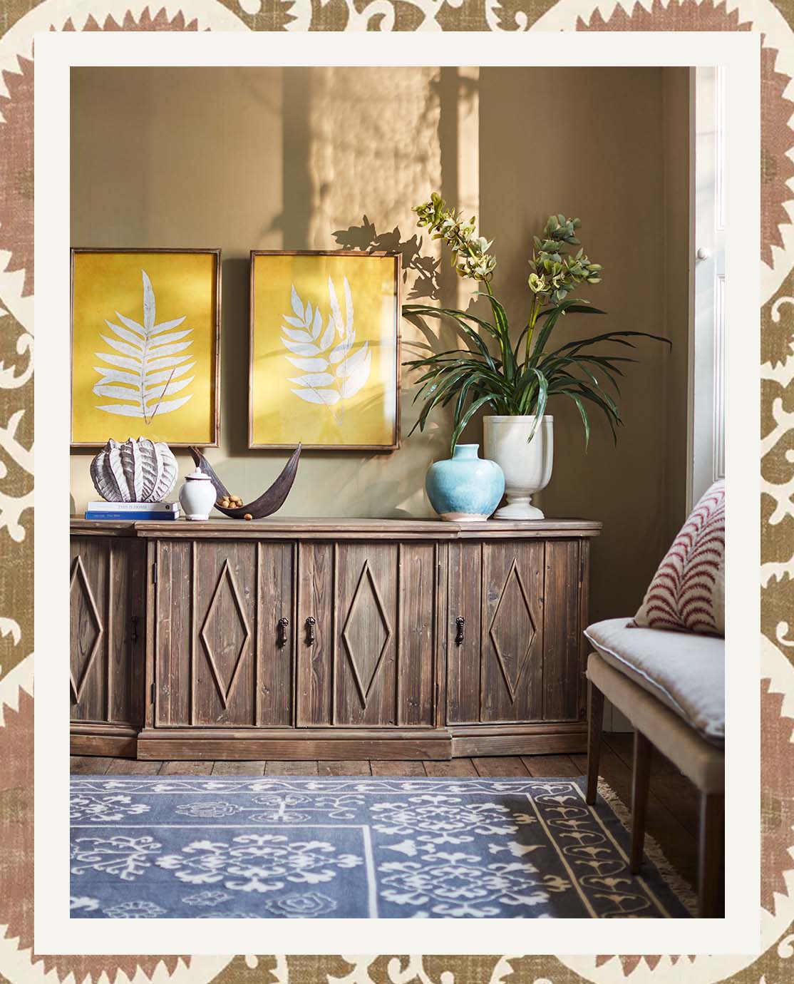 A sitting room setting with a blue printed rug, a linen upholstered bench, a wooden sideboard and yellow wall art