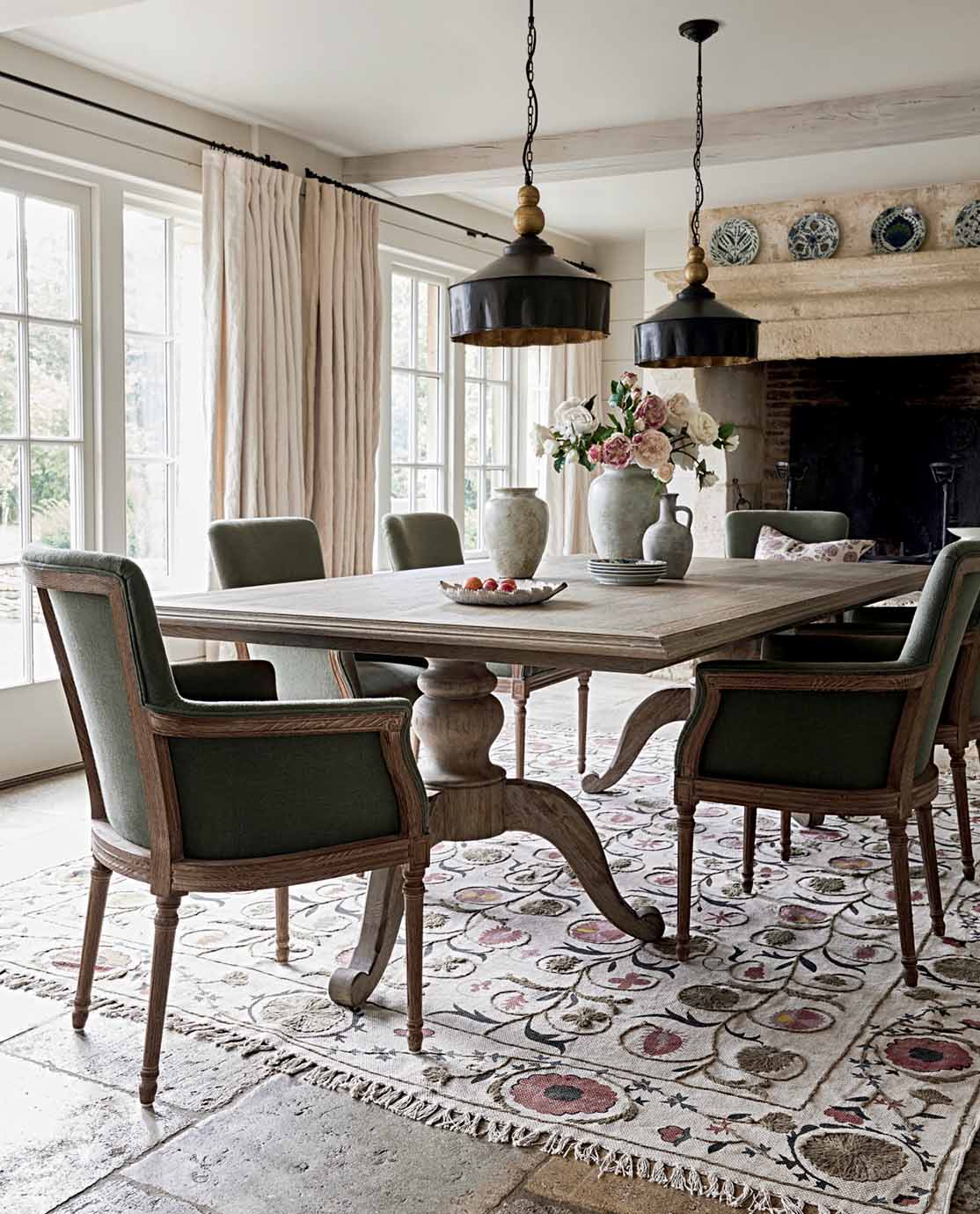 A set of green dining chairs with arms are arranged around a wooden dining table, which sits on a floral embroidered rug.