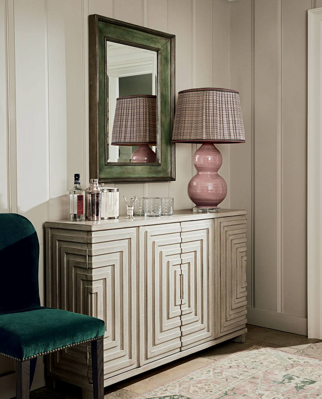 A Cubist-inspired cabinet is decorted with a pink lamp and glassware. A green velvet chair sits next to it.