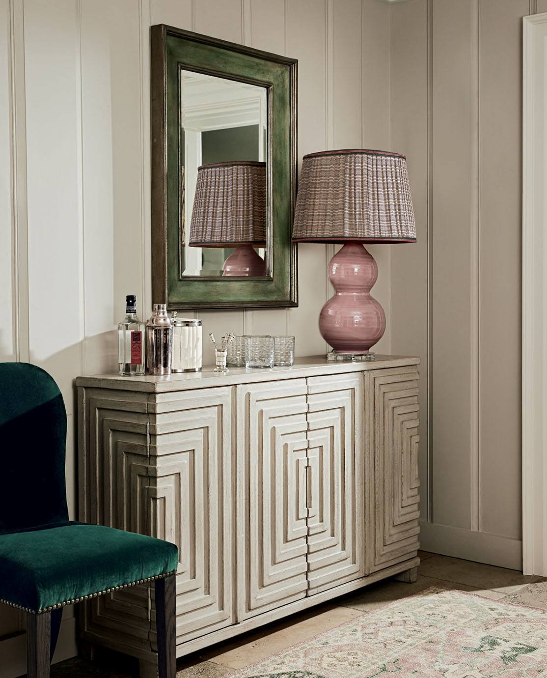 A Cubist-inspired cabinet is decorted with a pink lamp and glassware. A green velvet chair sits next to it.