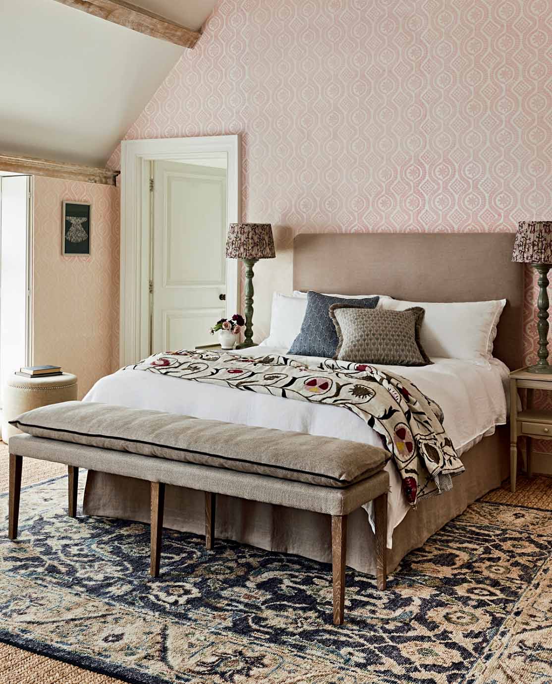 A country house bedroom with pink patterned wallpaper, a floral throw on the bed and a blue patterned rug.