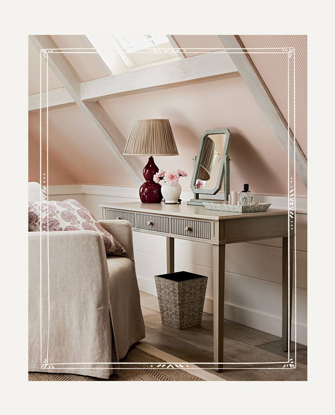 A armchair with a linen loose cover is pulled up to a wooden dressing table, which is decorated with a red lamp and a small faux shagreen mirror.