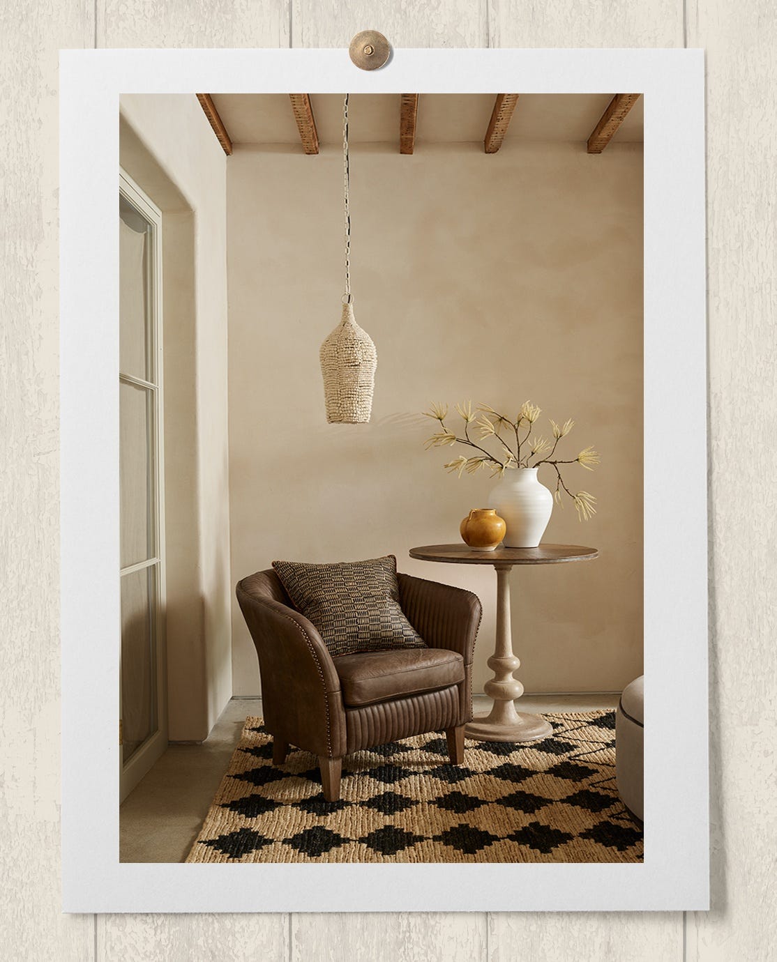 A pendant lampshade made from white, clay beads hangs above a brown leather armchair