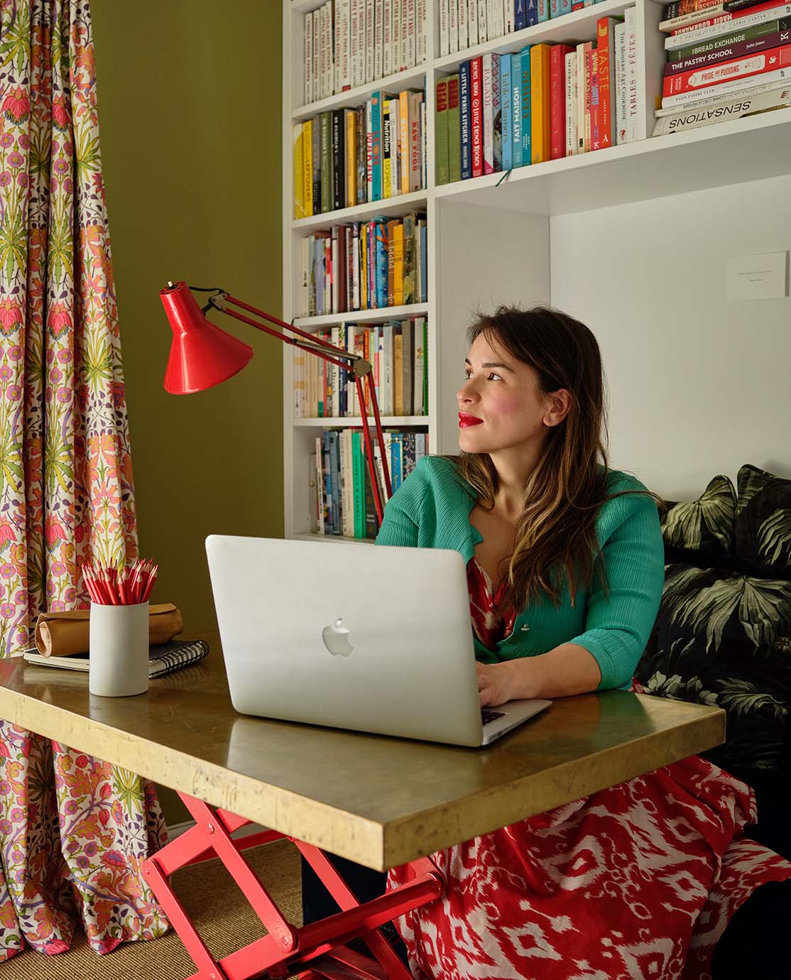 Rachel Khoo is wearing a green cardigan, sitting at a desk in front of an Apple Mac laptop. She is smiling and gazing off into the distance.