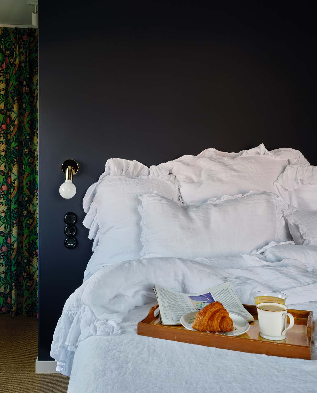 A bed dressed in white cotton sheets with frilly pillow covers. On top of the duvet is a breakfast tray holding a mug and a croissant.