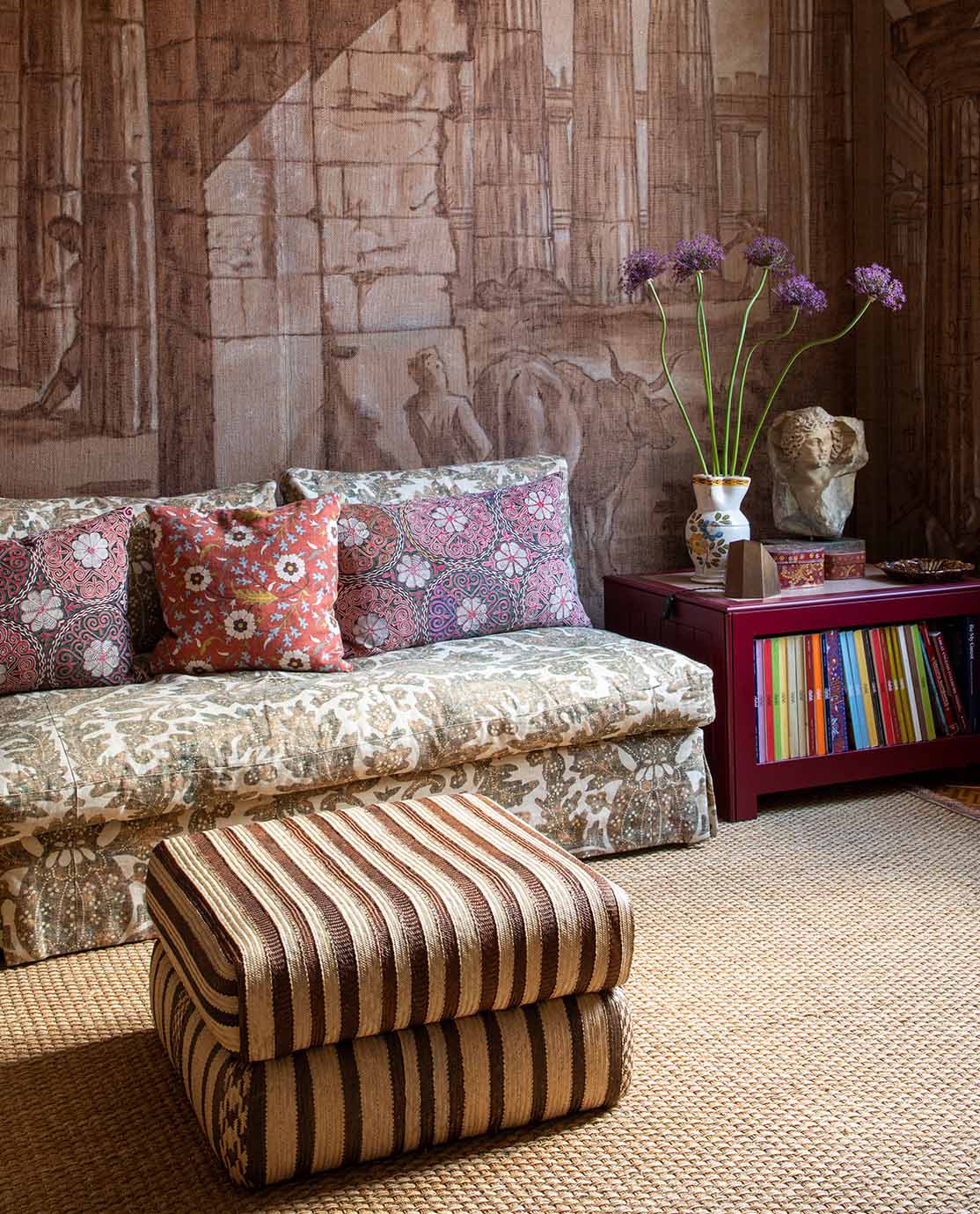 A green floral sofa decorated with pink and purple floral pillows and a striped ottoman sit in front of a mural of the Peastum ruins in Italy