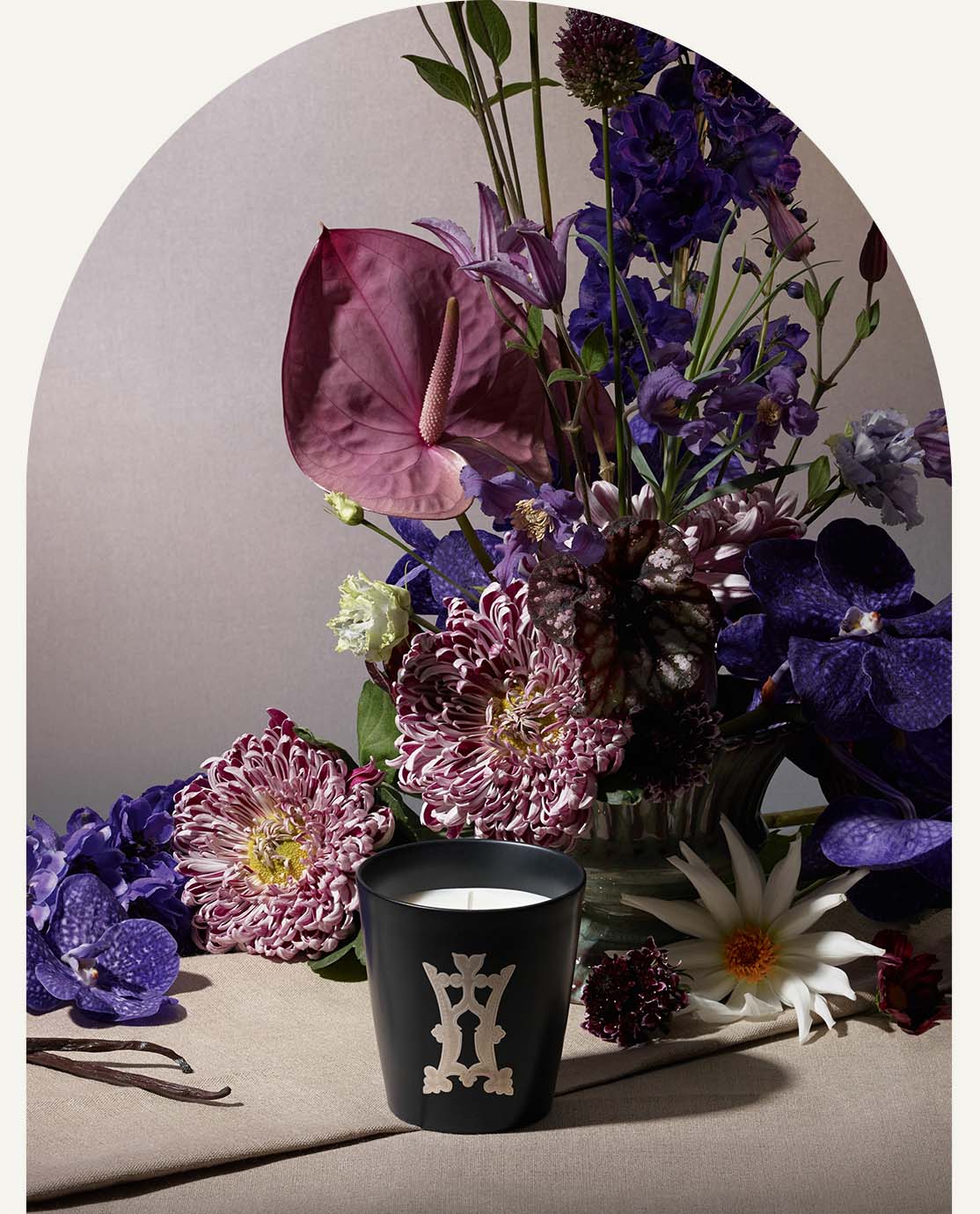 A candle in a black vessel sits in front of a bouquet of purple flowers