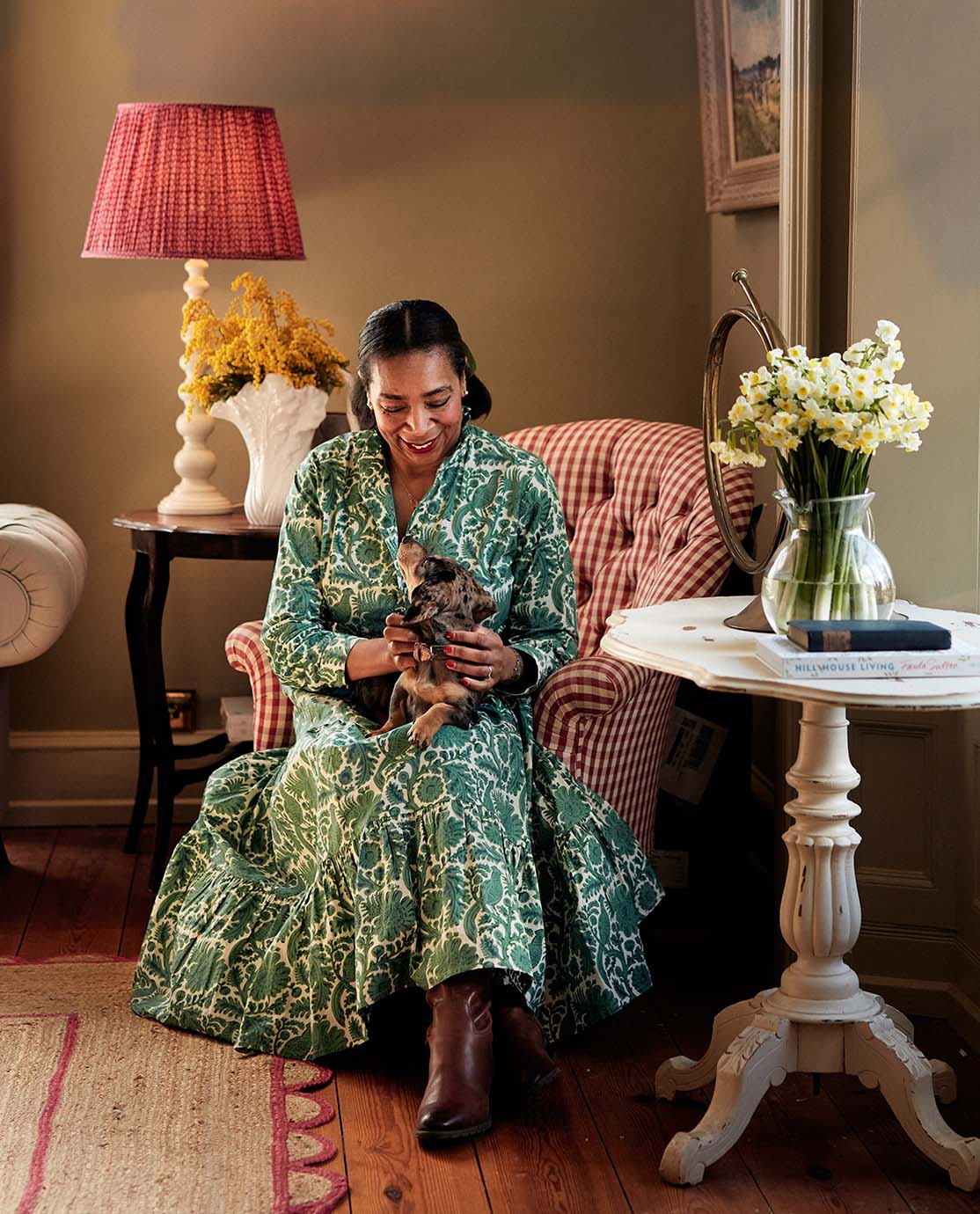 Paula Sutton is sitting in a red and white gingham armchair with a sausage dog on her lap.