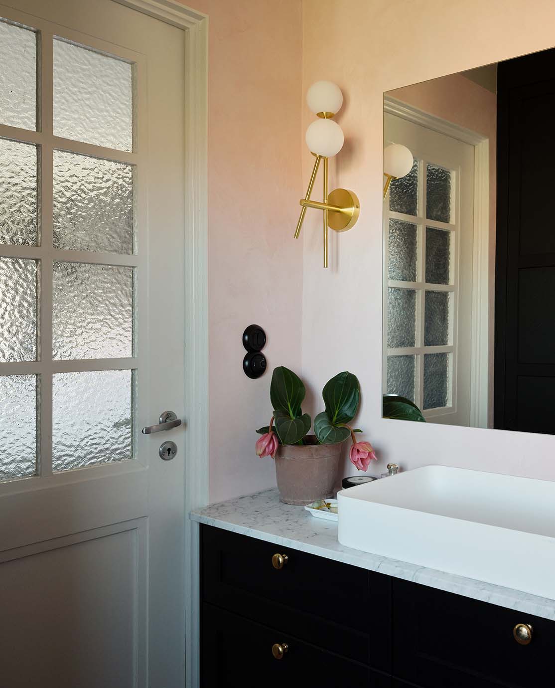 The corner of a light pink bathroom, with a large mirror and a navy cabinet. 