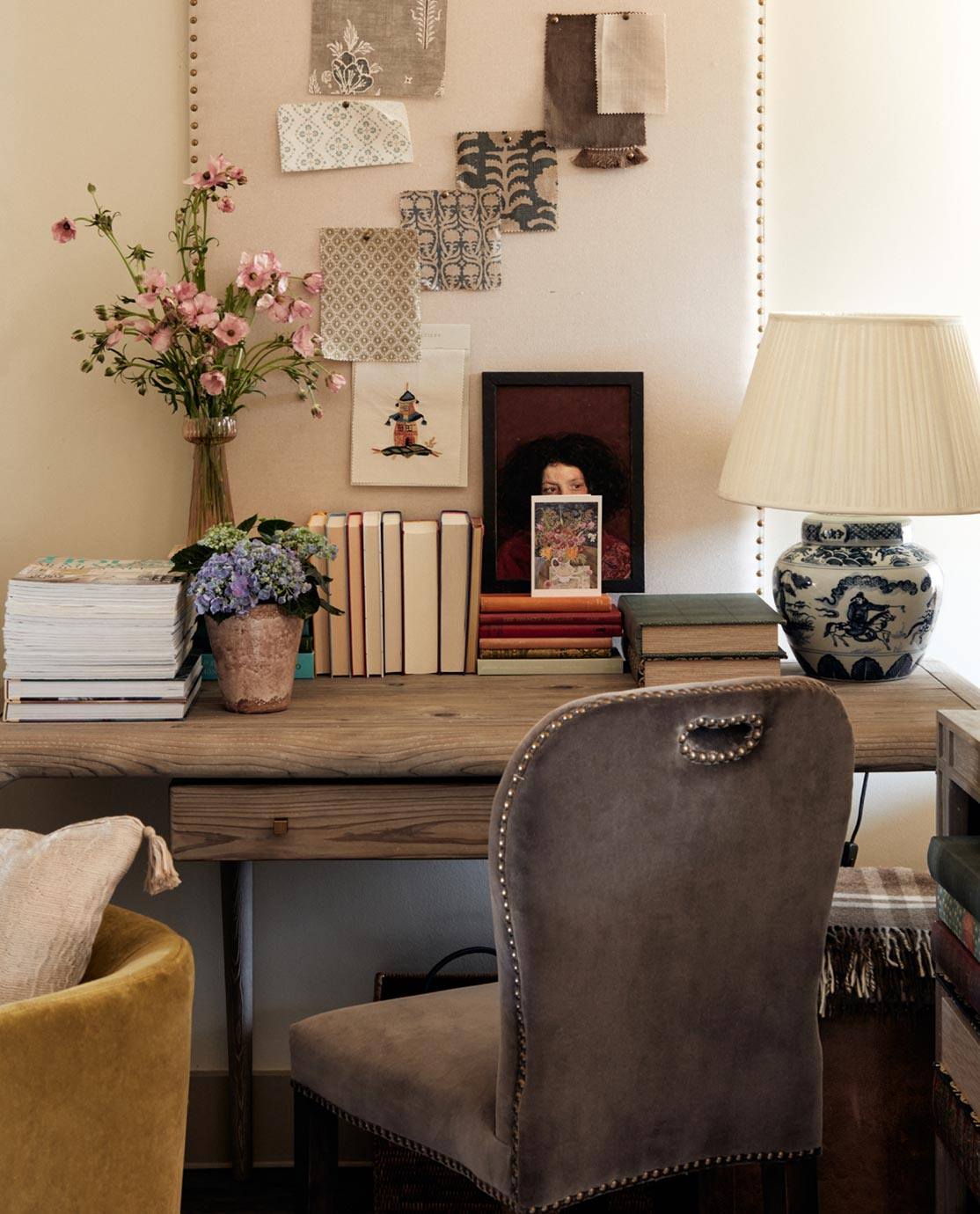 A velvet chair is pulled up at a wooden desk, which is decorated with books and a moodboard of patterned fabric swatches