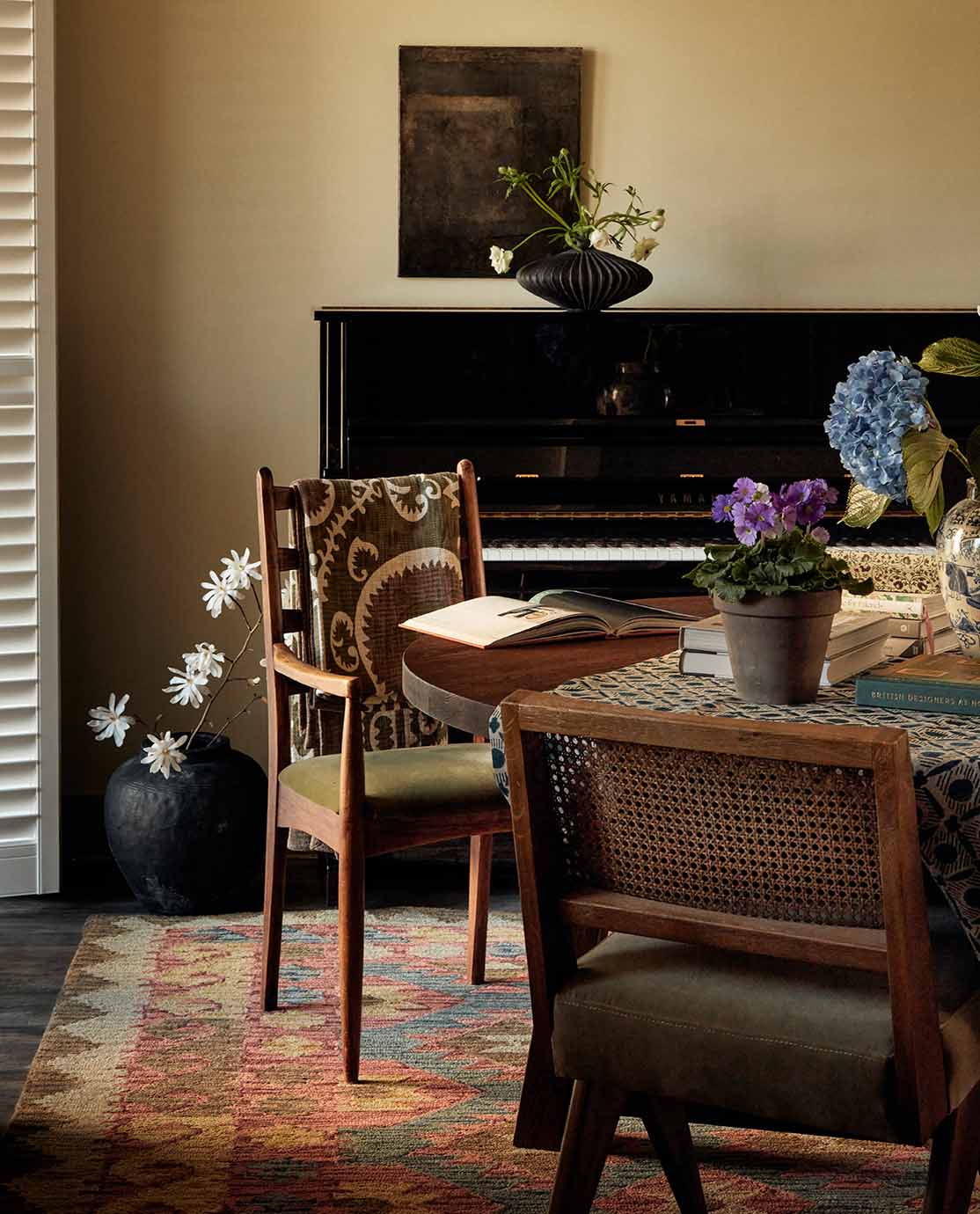 A dining room setting with a round wooden dining table, dining chairs upholstered in green velvet, and a black piano.