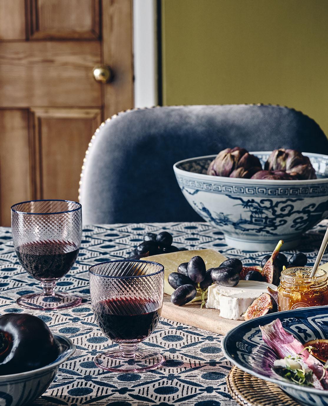 A dining table decorated with a blue patterned tablecloth, crockery and glassware.