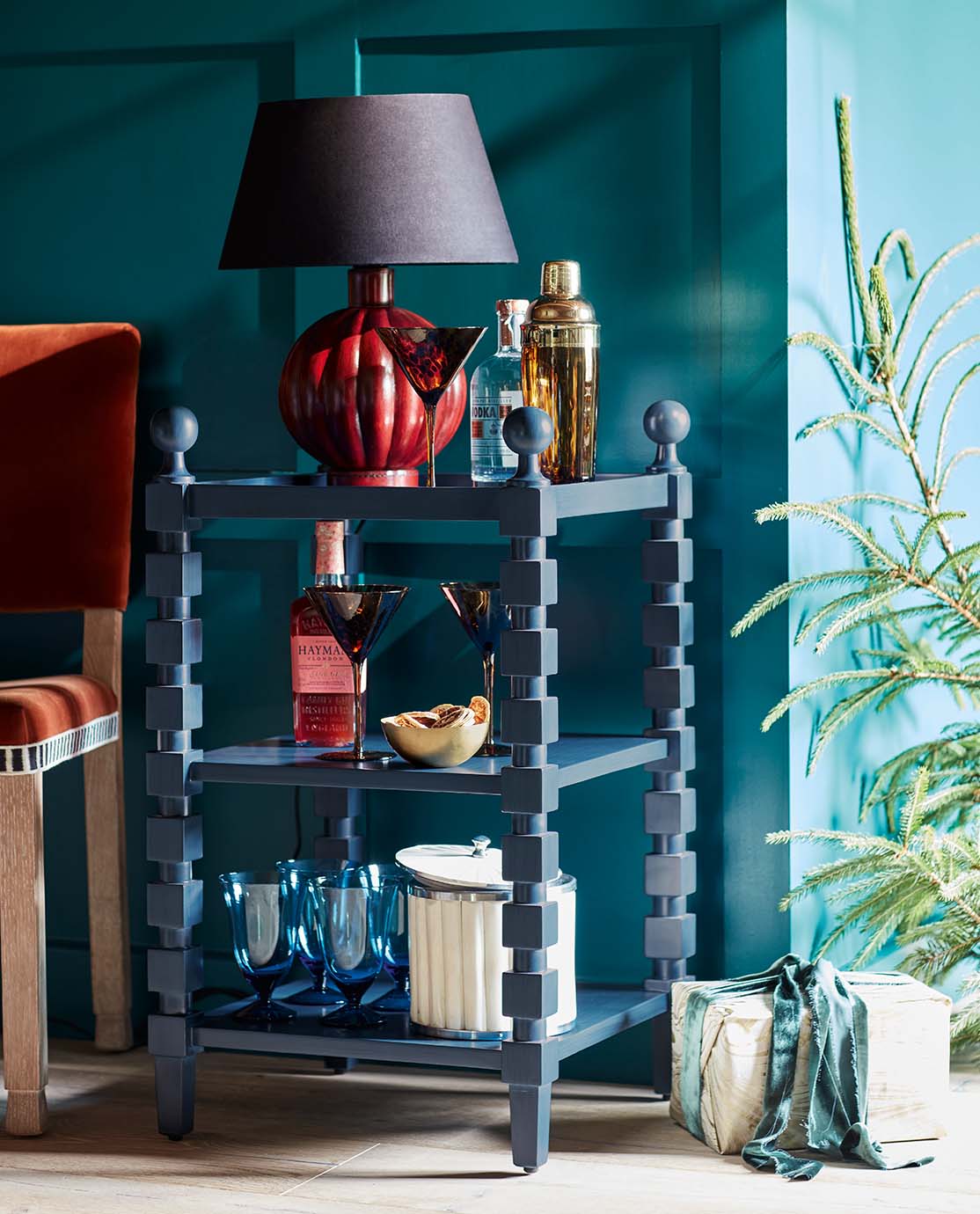 A blue side table featuring a red lamp, a cocktail shaker, tortoiseshell martini glassware and sapphire blue goblets