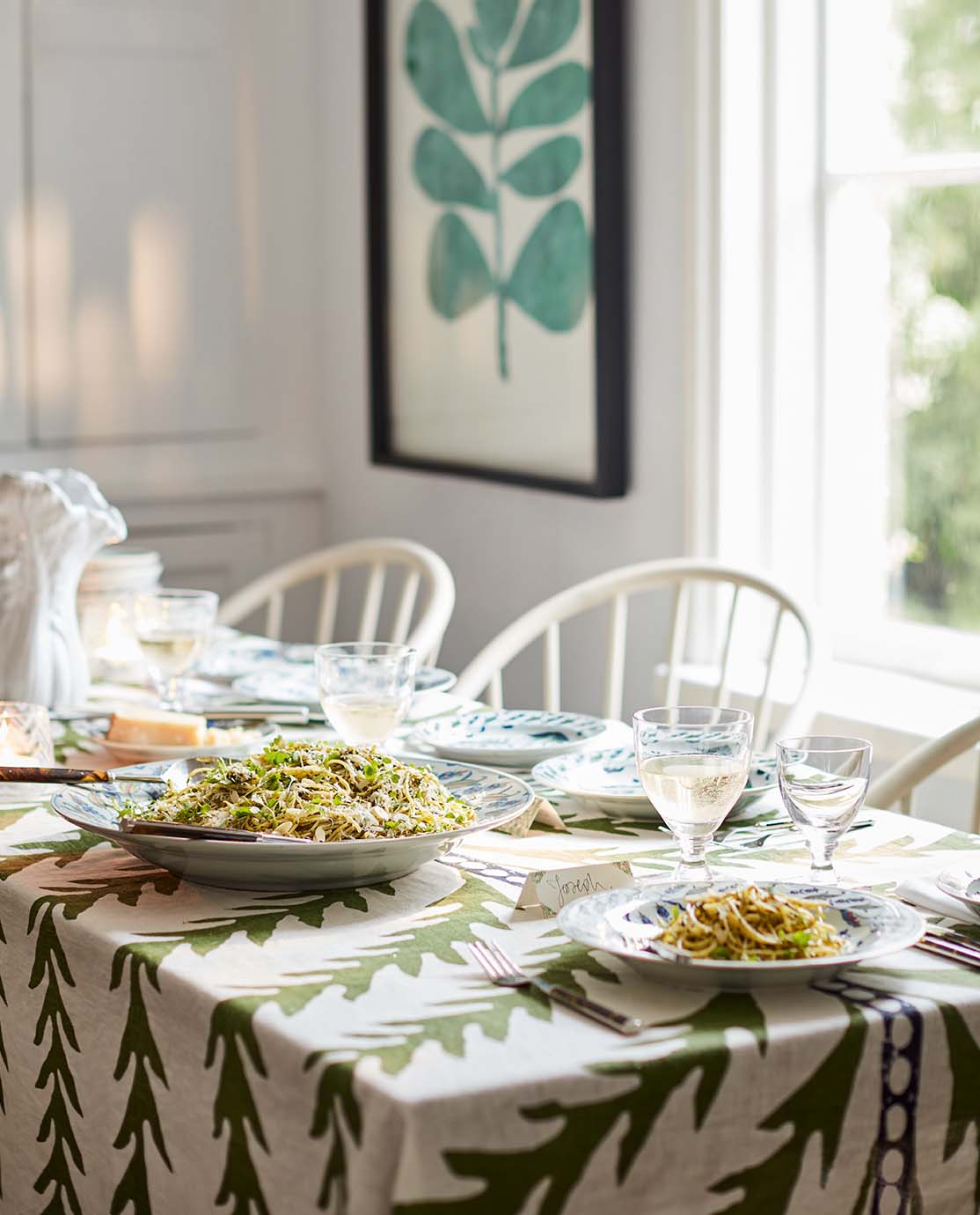 A table setting with a leaf-printed tablecloth, white dining chairs and patterned crockery