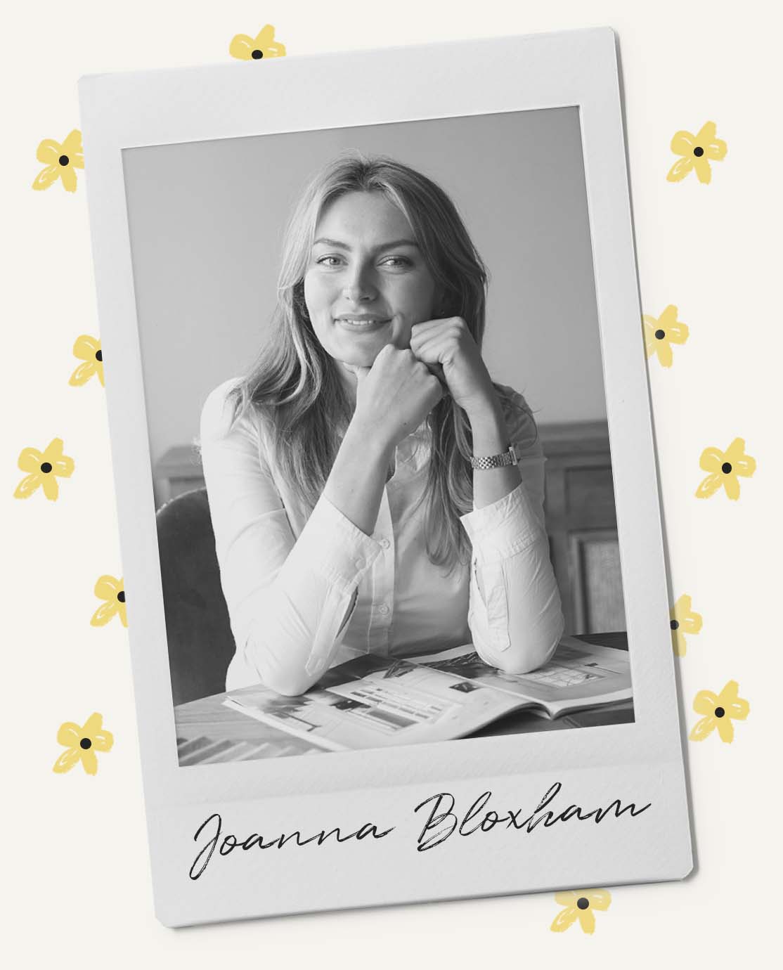 Joanna is wearing a white shirt and is sitting at a table leaning on her hands, smiling at the camera