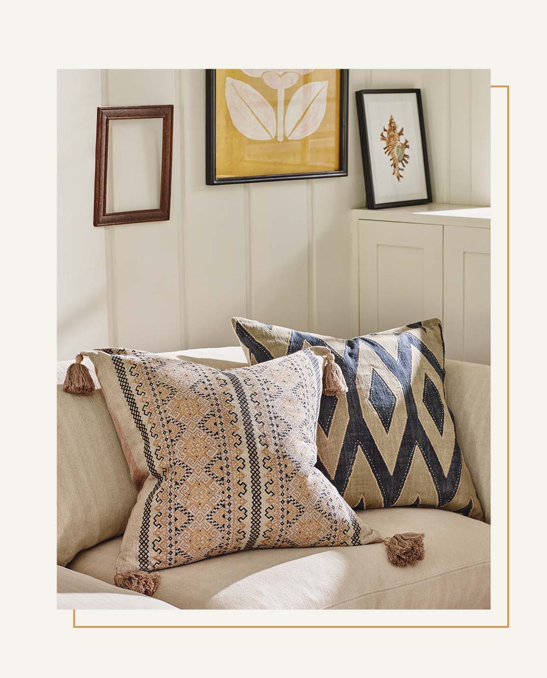 Two printed orange and blue pillows sit on a neutral-colored sofa. In the background, yellow and blue prints are hung on a white-paneled wall.