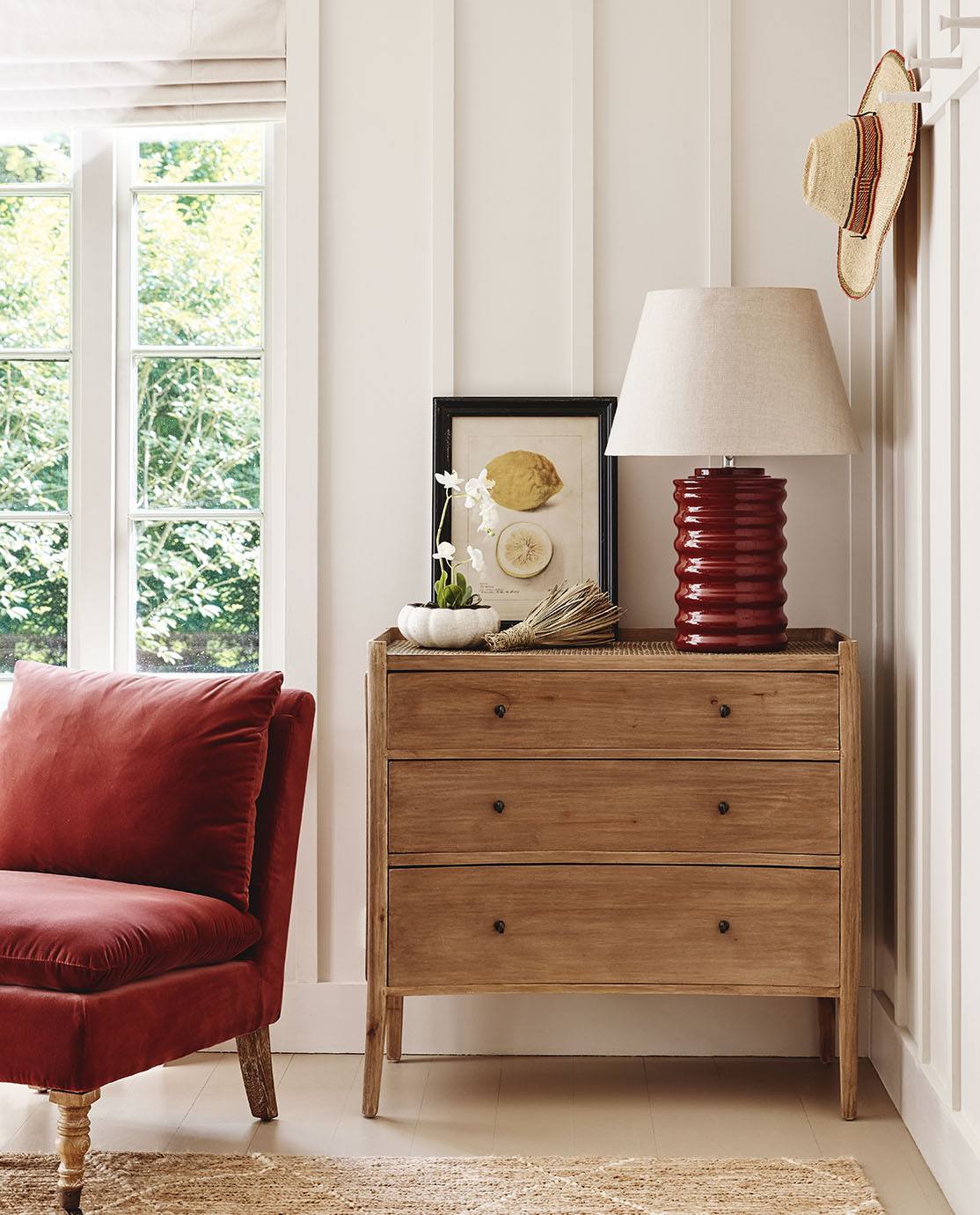 A rattan lamp is decorated with a red lampshade. It sits on a white nightstand. Above it hang two hats on wooden pegs.