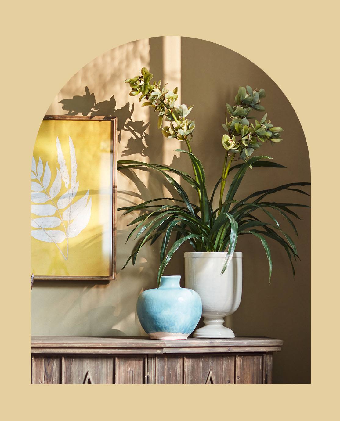 A large white vase holds a green plant. It is sitting on a wooden sideboard next to a blue pot