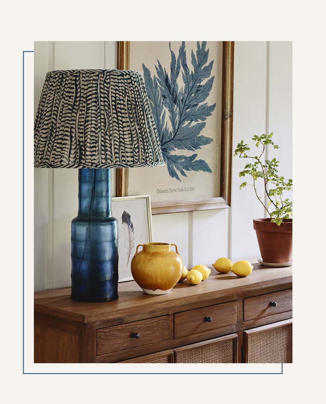 A blue lamp with a printed shade sit on a wooden sideboard, alongside a yellow ceramic pot, lemons and a terracotta plant pot.