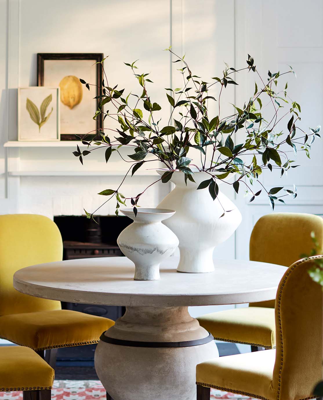 Two large white vases, one of which is holding foliage, sit on a round stone table surrounded by yellow velvet chairs.