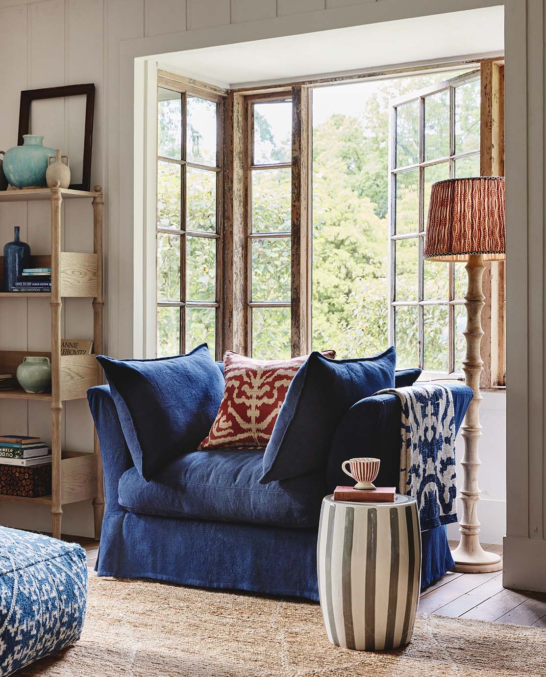 A cozy, cottage sitting room setting, featuring a navy blue loveseat, decorated with red pillows, a wooden lamp with a red lampshade and a ceramic striped stool.