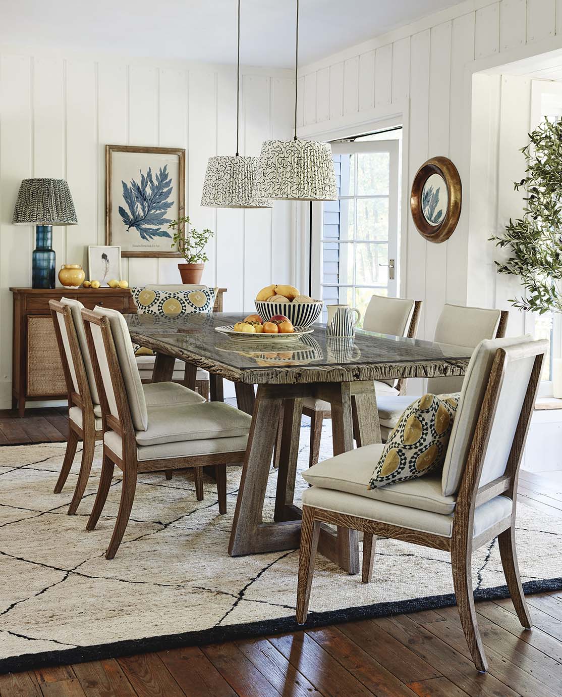 A light and airy dining room, with a wooden and glass dining table, white linen-covered chairs and printed lampshades.