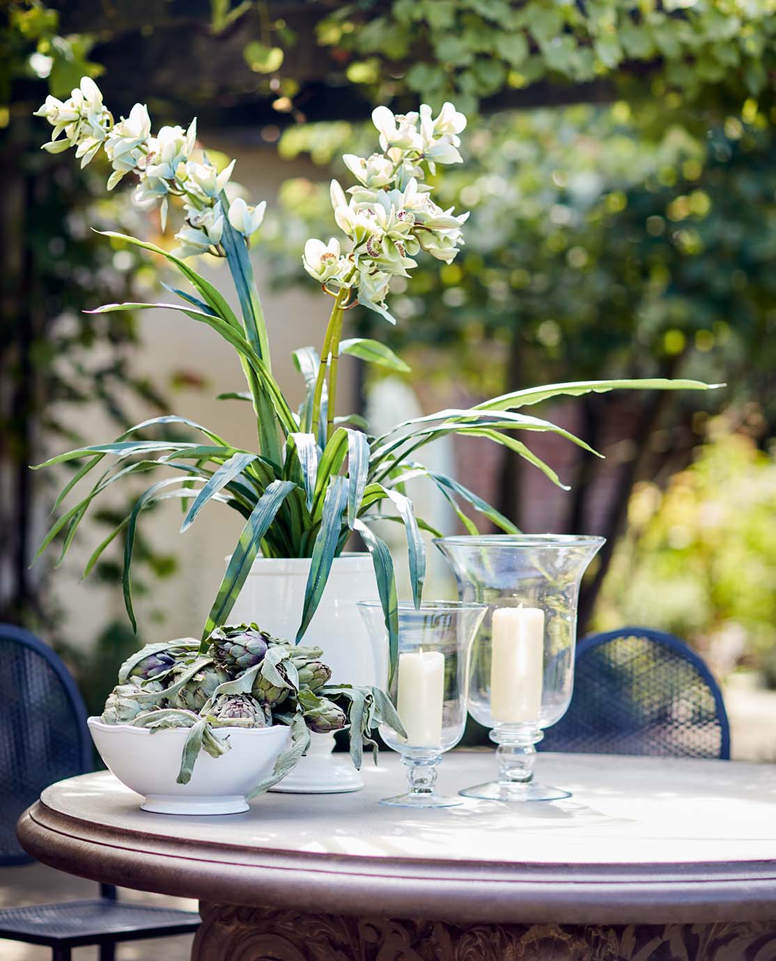 A white vase, holding a green plant, sits on a round outdoor dining table next to a white bowl of artichokes and two glass candle holders.