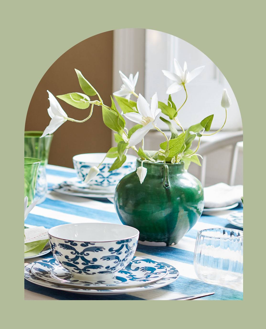 A green pot holding a stem of white flowers sits on a dining table, which is decorated with a blue-and-white striped tablecloth and floral crockery.