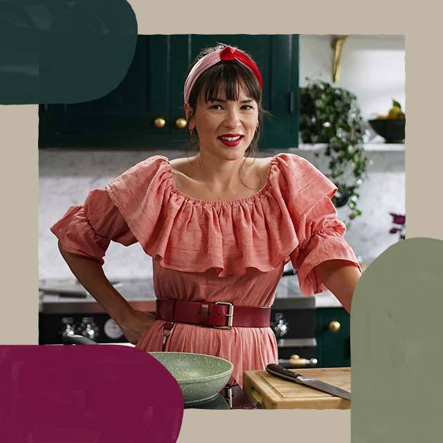 Rachel Khoo is wearing a pink ruffled off the shoulder dress with a red belt. She is standing in her kitchen smiling with her hand on her hip.