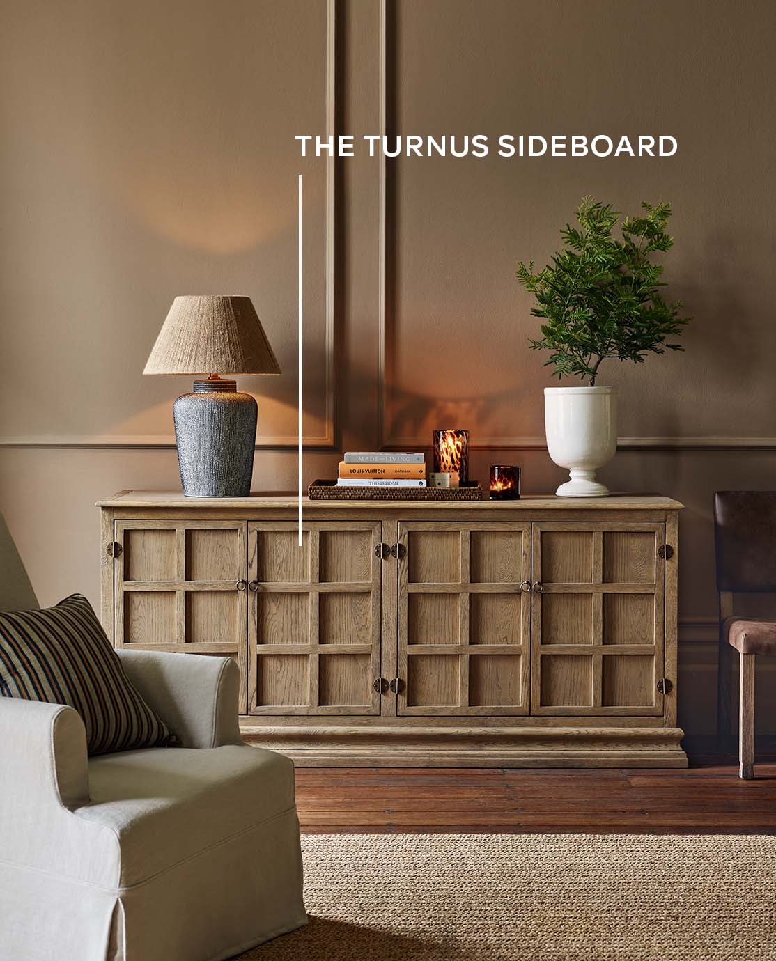 A wooden sideboard with a lamp, a vase and books in front of a white armchair.