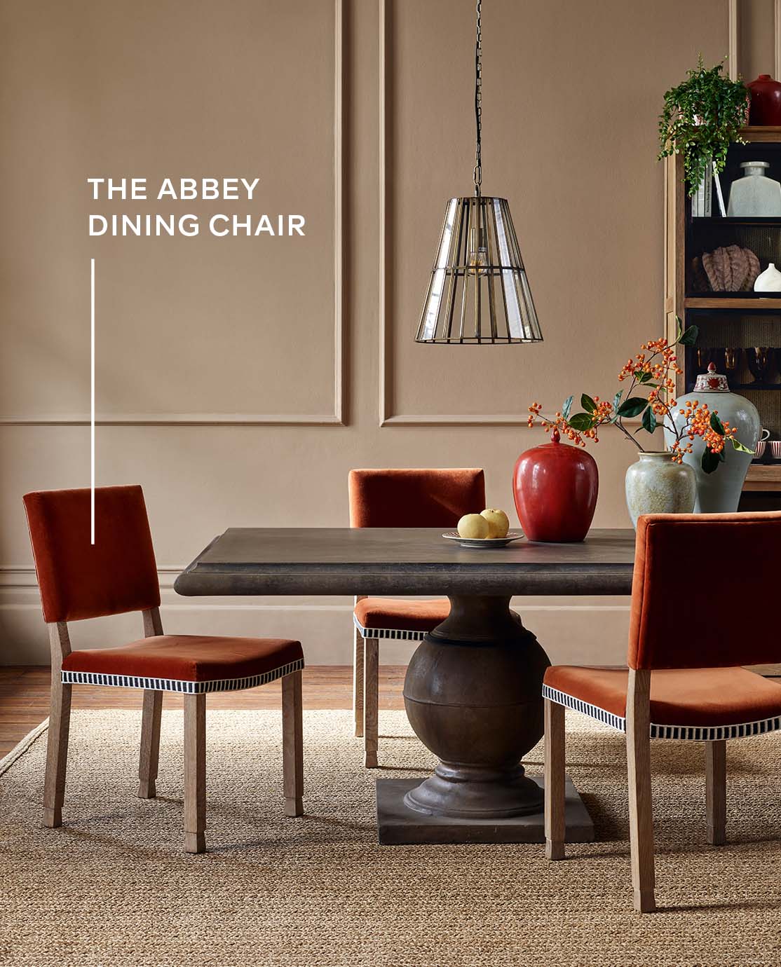 An autumnal dining room setting with a large wooden dining table, orange velvet dining chairs, red and blue vases and a large shelving unit