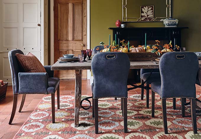 A sultry dining room with a large seasonal dining table in front of a dining table with blue chairs surrounding the table. The table sits on top of a multi-coloured rug.