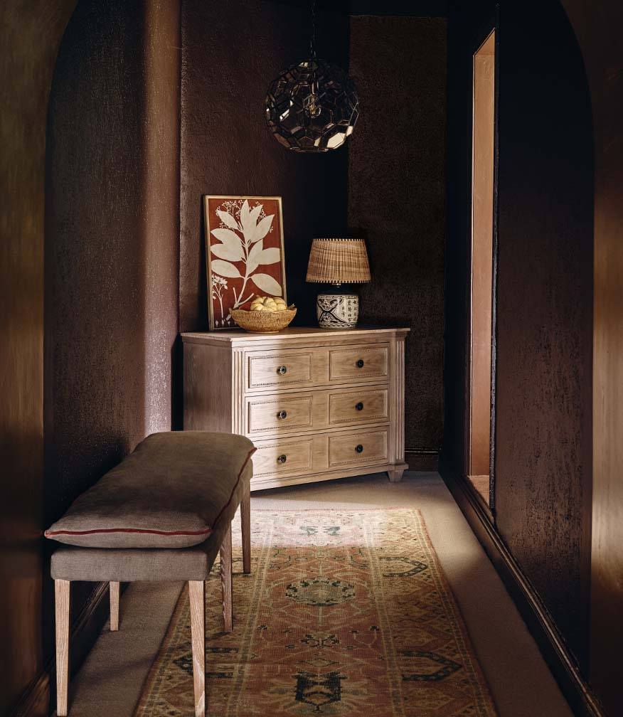 A dark hallway with a chest of drawers at the end of the hall, with an ornament, lamp and wall art on top. A bench sits in front of the hallway wall.